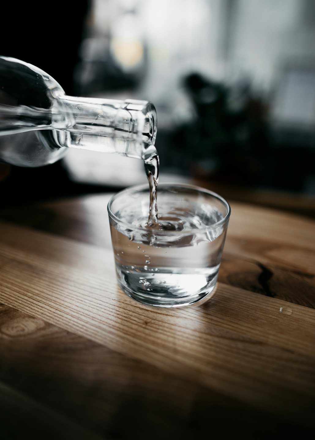Clear water pours from a glass bottle into a glass on a wooden table.